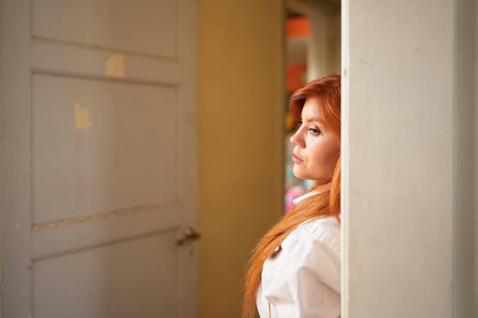 A woman with red hair leans against a doorframe in an indoor setting, looking pensive.