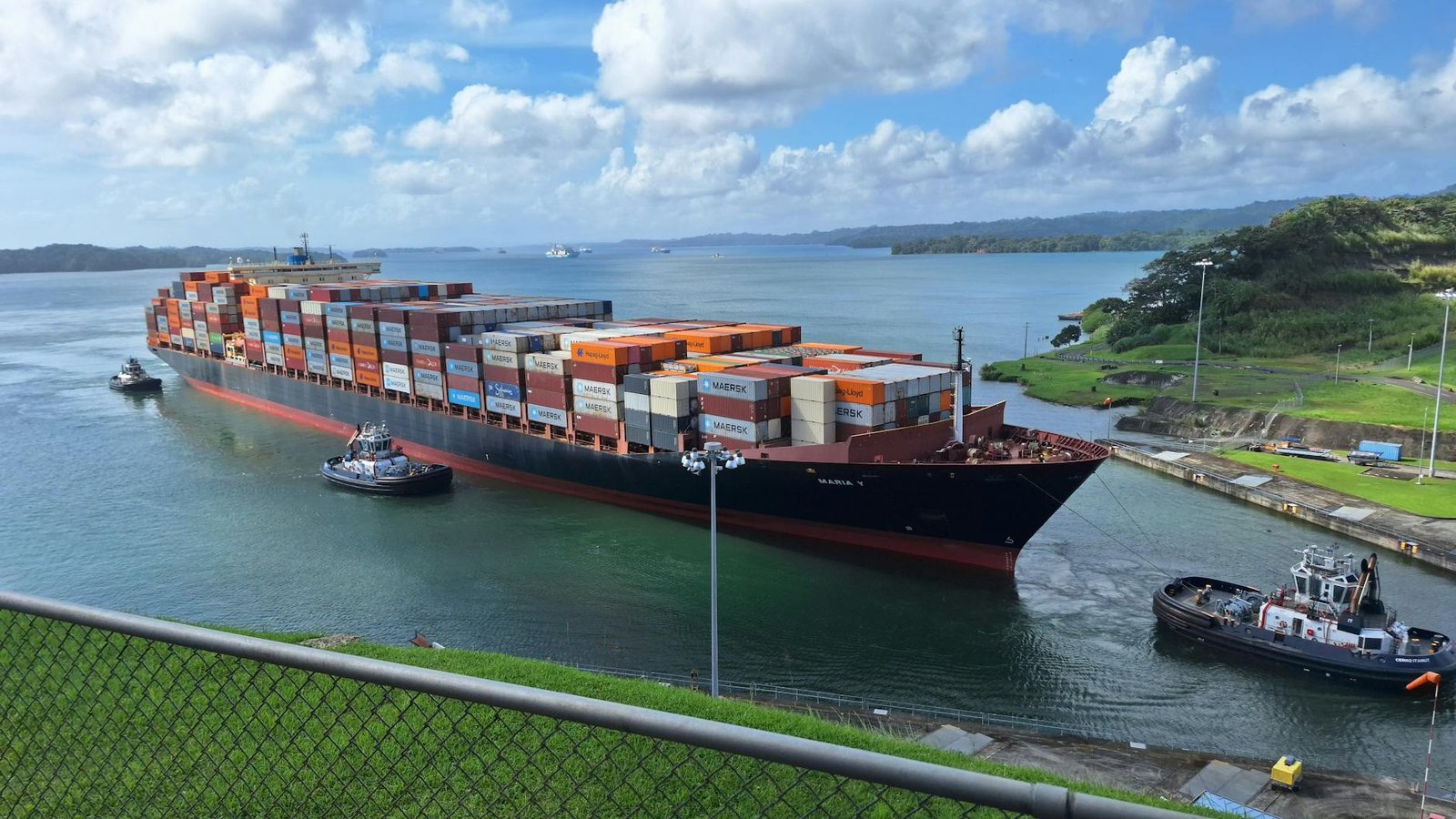 Large container ship maneuvered by tugboats through the Panama Canal under a clear sky.